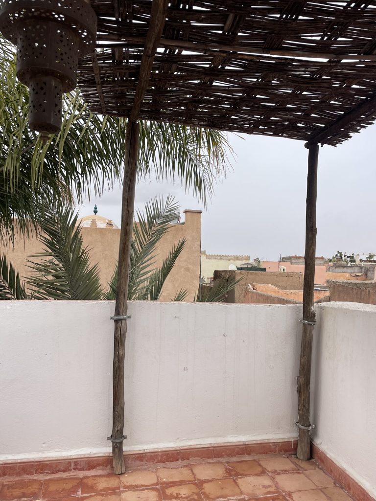 Terrasse sur le toit d’un riad à Marrakech avec pergola en bois, palmiers et vue sur les toits de la médina